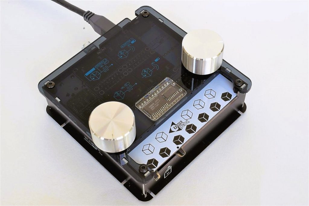 Assembled DAFM synth in a black acrylic case with silver knobs, a white panel with cube graphics and a backlit display